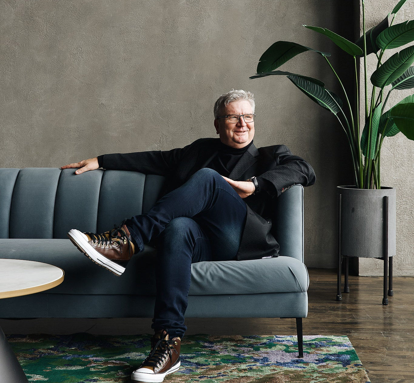 Studio 321B Founder Anthony Hellman sitting on a blue sofa, cross legged. Anthony is wearing a black turtleneck and black blazer with blue jeans. On the floor we have the Sustainability rug (green / gold / blue splotched rug)