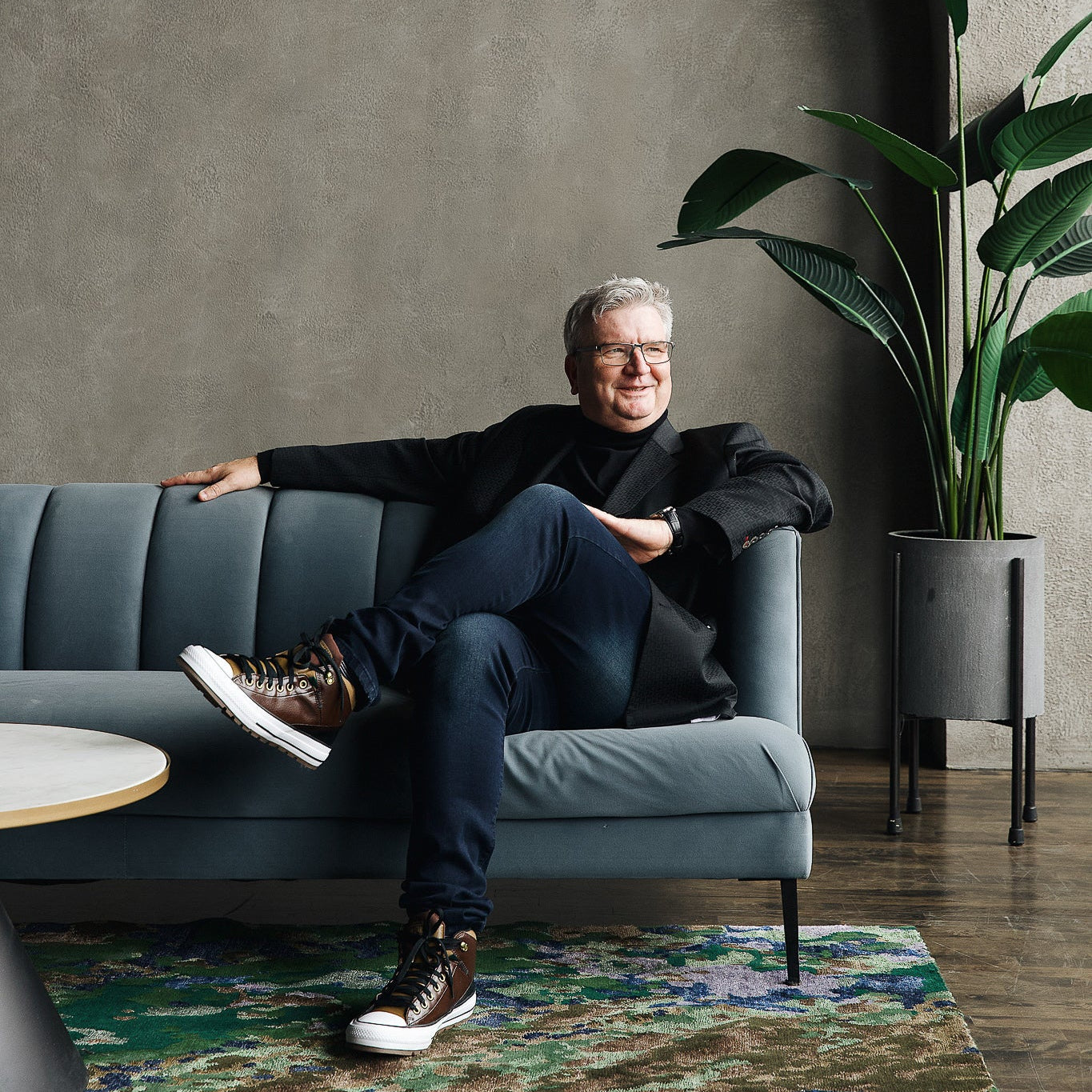 Studio 321B Founder Anthony Hellman sitting on a blue sofa, cross legged. Anthony is wearing a black turtleneck and black blazer with blue jeans. On the floor we have the Sustainability rug (green / gold / blue splotched rug)