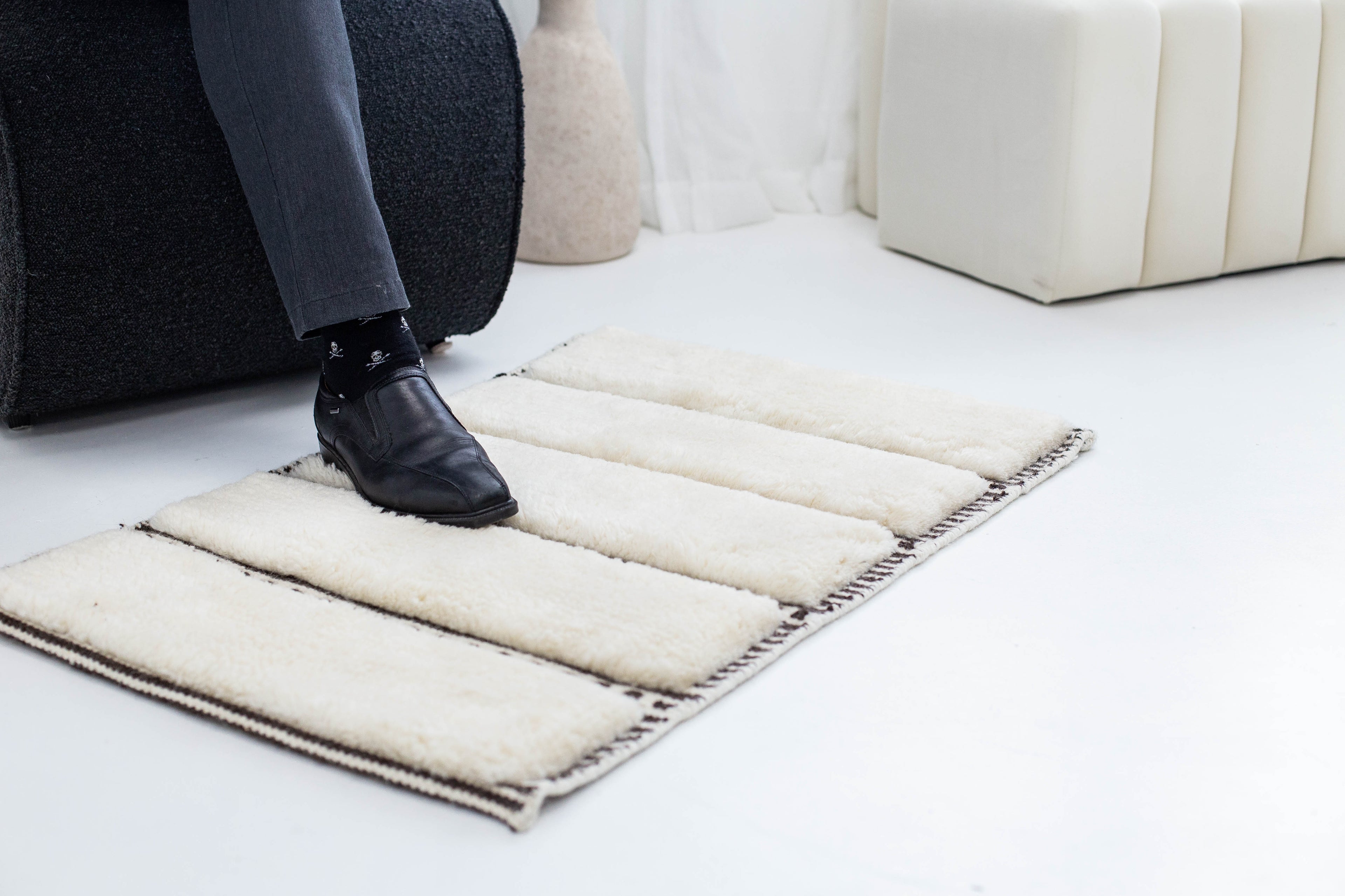Cream wool rug with dark stitched border, shown under a black leather shoe in a modern interior setting. Created by Studio321B rug studio Toronto, Ontario.